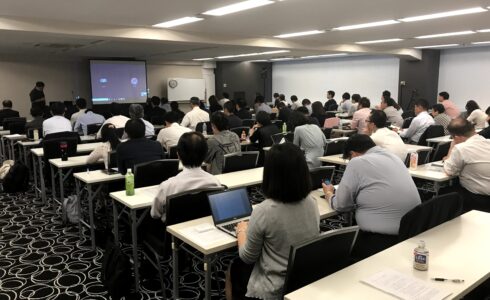 会議室で行われているセミナーの様子。多くの参加者がノートパソコンや資料を使いながら講演を聞いている。前方にはスクリーンが設置され、プレゼンテーションが行われており、講師がスクリーン横に立ち、台湾有名ブロガー林氏碧とオンラインで対話をしている。